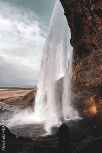 Vertical Wide Angle View of Seljalandsfoss Waterfall in Iceland from behind waterfall  with a silhouette of a woman in a fur hood jacket