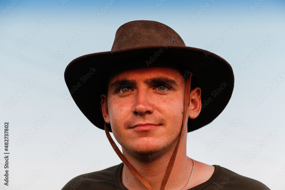 Young man smiling farmer in cowboy hat at field against blue sky. Portrait of millennial man standing on nature background, outdoors. Rancher on farmland. Happy summer. Closeup.  Countryman
