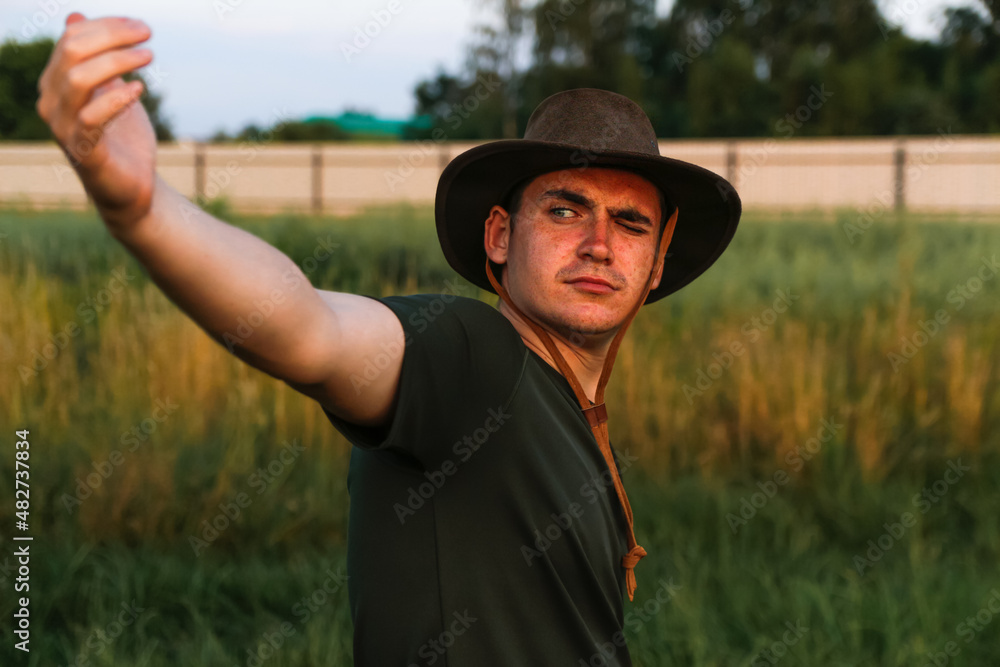 Young man serious in cowboy hat showing flying hands against green grass. Male on blue sky background, outdoors in meadow. Agriculture concept.  Wow and fun. Close-up. Hero wings, plane. Closed eye
