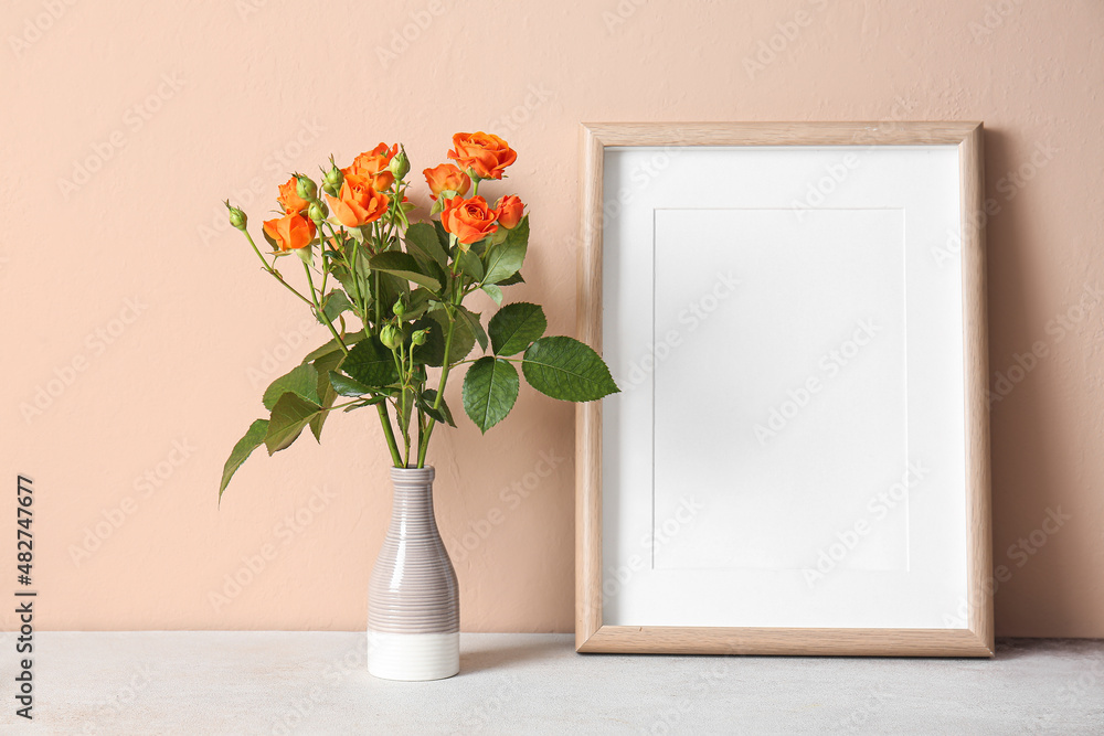 Vase with beautiful orange roses and blank photo frame on table against beige wall Vase with beautiful orange roses and blank photo frame on table against beige wall