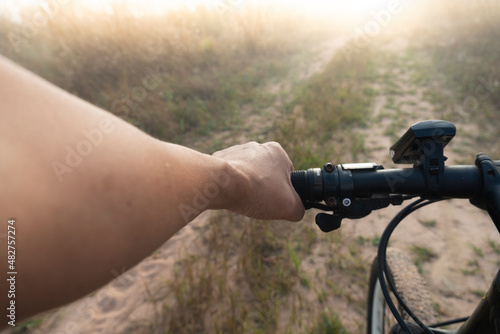 Cyclist's hand holds the handlebar of a bicycle and rides it on a dirt road. Golden yellow fields with the light shining and the mist in front of them faded.