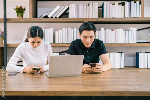 Young Asian male and female working together on the wooden table with laptop, looking at their mobile phones with a bookshelf in the background.