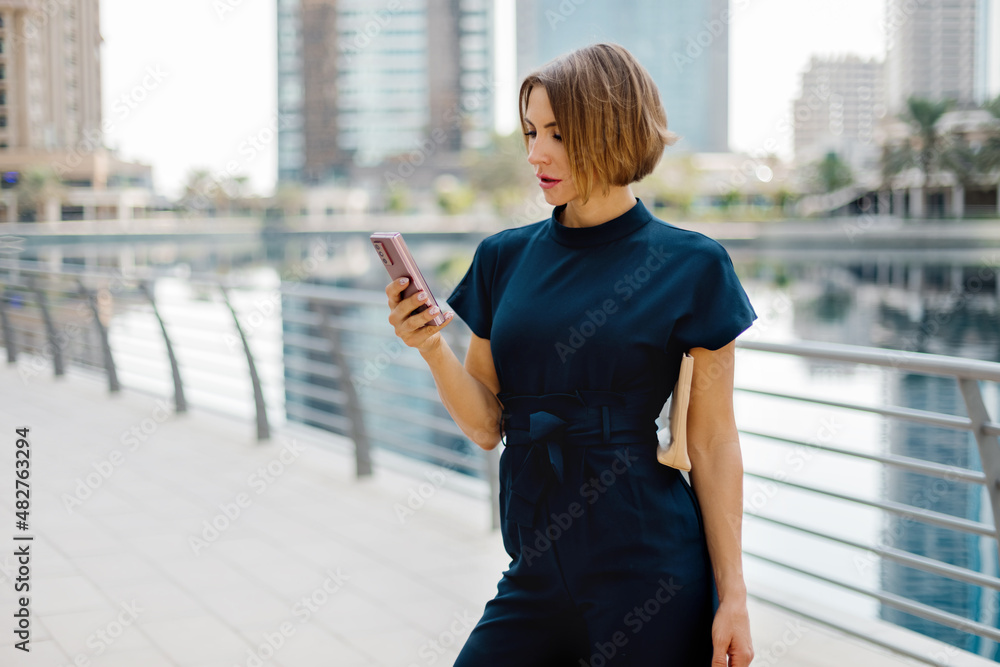 Fototapeta premium An attractive young woman using a smartphone while walking along the embankment