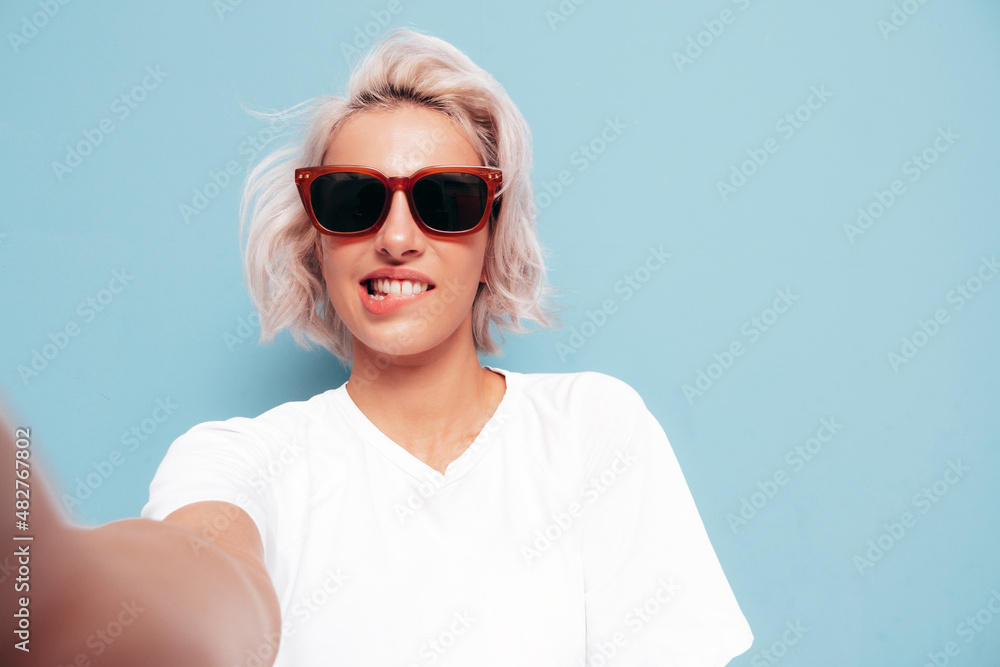 Young beautiful smiling female in trendy summer clothes. Sexy carefree woman posing near blue wall in studio. Positive blond model having fun in sunglasses. Cheerful and happy. Taking Pov selfie Young beautiful smiling female in trendy summer clothes. Sexy carefree woman posing near blue wall in studio. Positive blond model having fun in sunglasses. Cheerful and happy. Taking Pov selfie