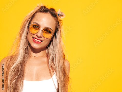 Portrait of young beautiful smiling blond female in trendy summer clothes. Sexy carefree woman posing near yellow wall in studio. Positive model having fun indoors. Cheerful and happy in sunglasses