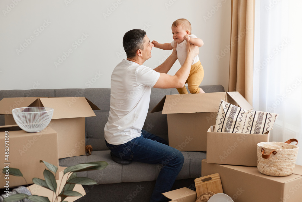 Side view portrait of man wearing white casual T-shirt and jeans, sitting on sofa with infant daughter, unpacking boxes after relocating, playing with child, pulling kid into cardboard box. Side view portrait of man wearing white casual T-shirt and jeans, sitting on sofa with infant daughter, unpacking boxes after relocating, playing with child, pulling kid into cardboard box.
