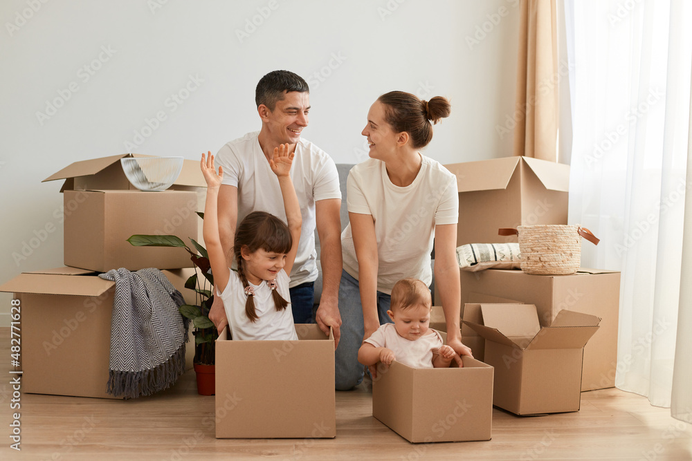 Horizontal shot of happy family relocating to a new flat, riding their children in cardboard boxes, having faun together, celebrating relocation to a new apartment. Horizontal shot of happy family relocating to a new flat, riding their children in cardboard boxes, having faun together, celebrating relocation to a new apartment.