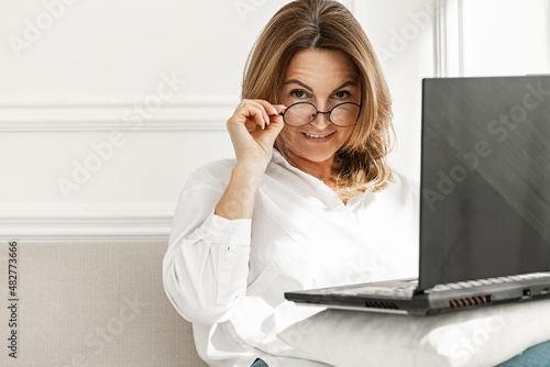 Portrait of an attractive middle-aged woman in stylish glasses. Beautiful positive smiling woman in white cotton shirt sitting on sofa with laptop. Copy space