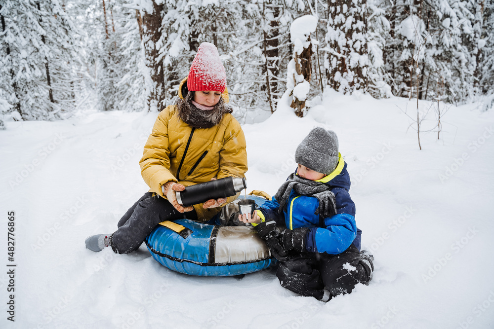 Mother and son sit in the snow, drinking tea. Winter outdoor recreation. Treat each other to hot tea. A family adventure, a walk in the park in winter.