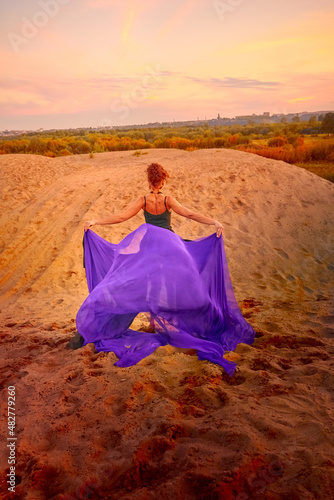 Beautiful girl with black dress and violet fluttering cloth dancing on the sand dune. Model or dancer posing in nature landscape