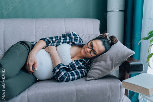 Dark-haired pregnant woman in and plaid shirt lying on comfortable grey couch in modern living room. Female feel pain or discomfort and hug belly. Contractions, braxton hicks  photo