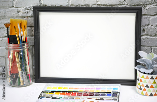 Black landscape frame beside a small houseplant and watercolor supplies, painting or artwork display mockup in front of vintage white brick wall.