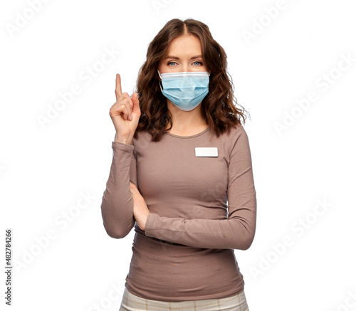 sale, shopping and pandemic concept - happy female shop assistant in medical mask with name tag pointing finger up over white background