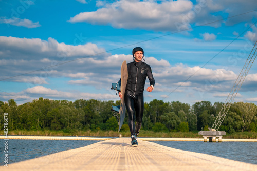 Confident man in wetsuit with wakeboard walking on floating bridge after training