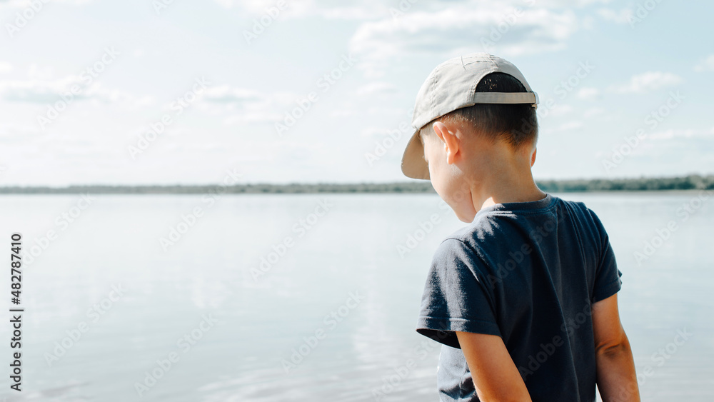 Back view of lonely little caucasian boy in cap standing on shore of lake on summer sunny day, copy space. Rear view pensive child in nature, outdoors