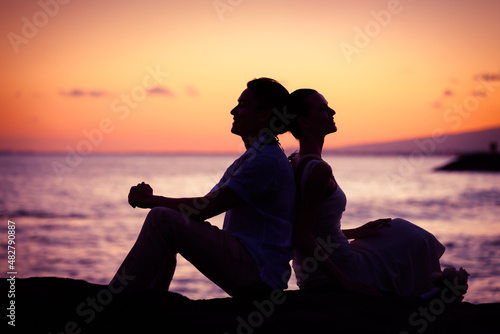 Happy couple silhouette sitting on beach back to back looking up to the sky