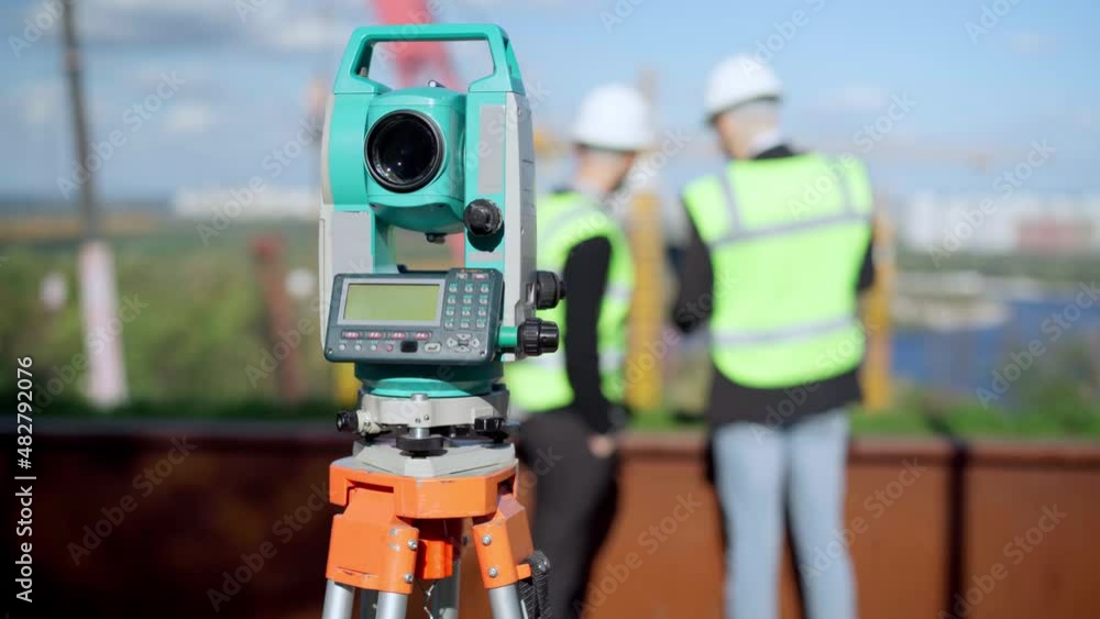 Close-up theodolite with two blurred men talking at background in sunshine. Angular measurement surveying instrument outdoors at construction site with male engineers discussing project Close-up theodolite with two blurred men talking at background in sunshine. Angular measurement surveying instrument outdoors at construction site with male engineers discussing project