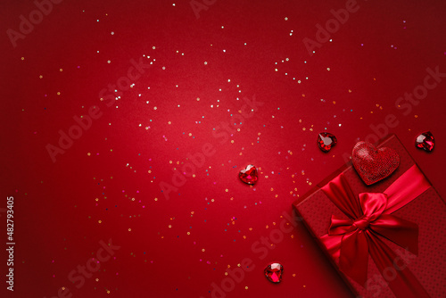 Valentine's Day gift in a red box tied with a red ribbon and red crystals, heart-shaped rubies, rhinestones, glitter on a red background.