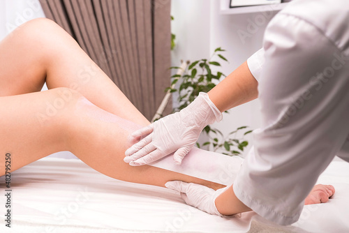 Close up photo of woman patient legs on the massage cosmetic table and cosmetologist with gloves making waxing procedures using warm cartridge wax in a beauty salon.