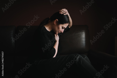 Frustrated woman dressed in black sitting on black sofa with hands on head experiencing negative emotions. Depression, grief, stress, loneliness, loss, fear, dissipation, sacrifice. Mental health