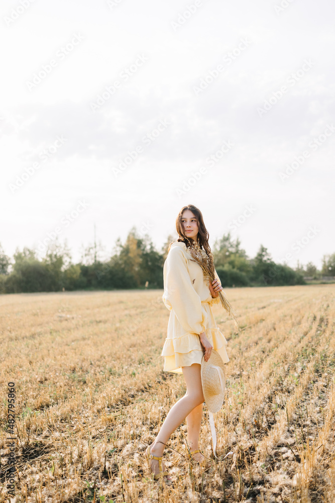 Happy young girl running through a yellow field on a summer day. girl in yellow dress and straw hat with a bouquet of wheat. sheared golden wheat spikelets. Agricultural texture Happy young girl running through a yellow field on a summer day. girl in yellow dress and straw hat with a bouquet of wheat. sheared golden wheat spikelets. Agricultural texture