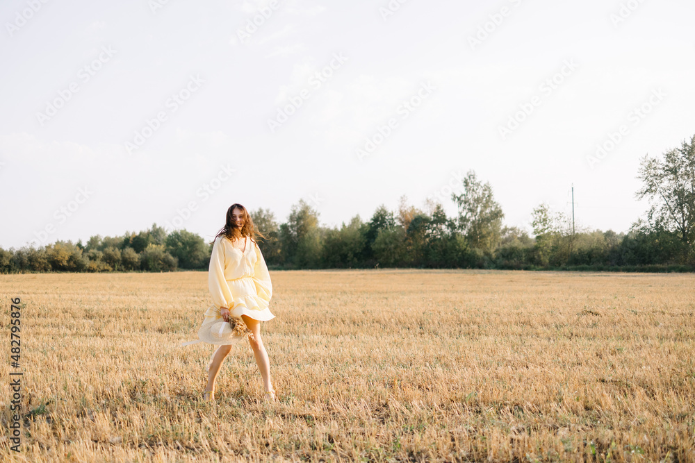 Happy young girl running through a yellow field on a summer day. girl in yellow dress and straw hat with a bouquet of wheat. sheared golden wheat spikelets. Agricultural texture