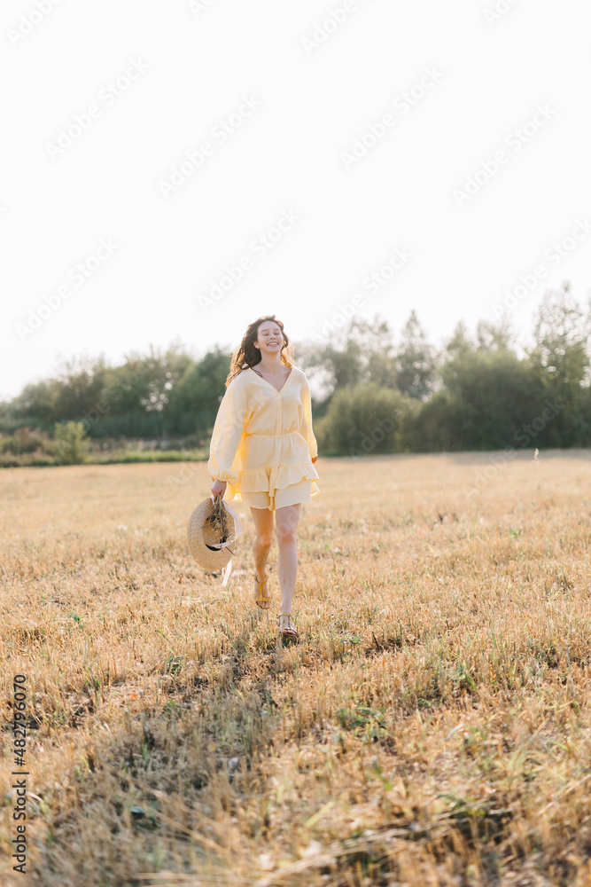 Caucasian young brunette walking through a wheat field. girl in yellow summer dress and straw hat. sheared golden ears of wheat, rye. Close-up portrait of a beautiful girl with brown eyes. 