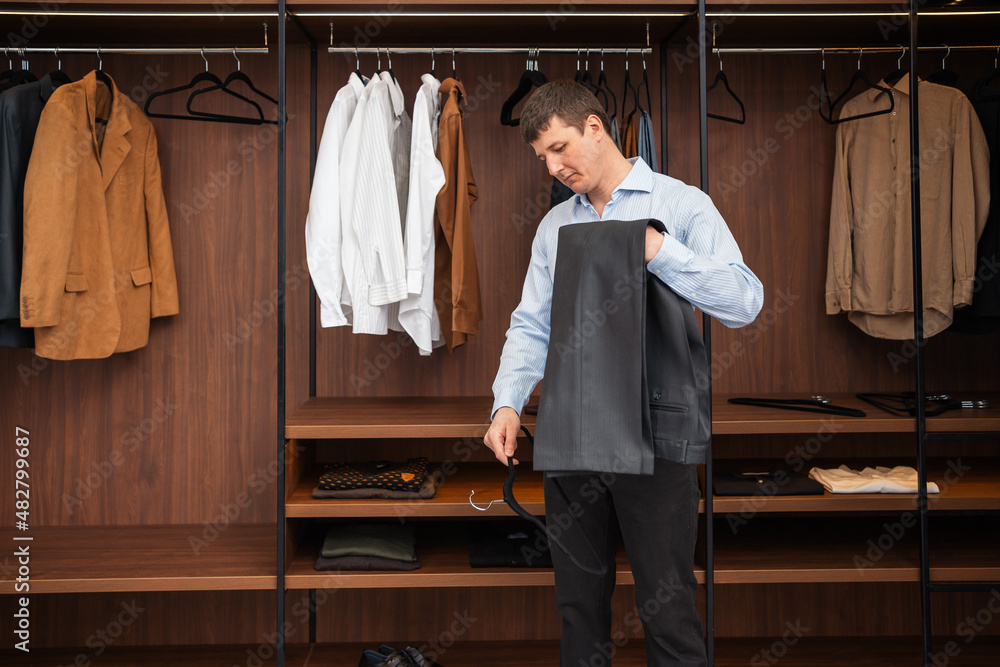 Young European man with dark hair brings wardrobe order puts everything in its place. Closet organized. Man chooses clothes. Capsule wardrobe for men.