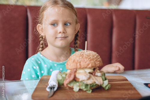 Portrait of a little girl with blond hair and blue eyes sitting next to the wooden tray with a huge tasty burger. Fast food concept.