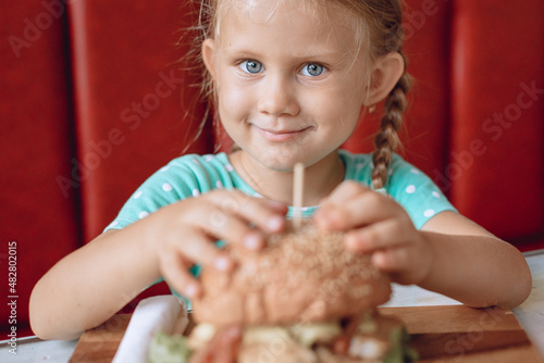 Little funny smiling child with blond hair is looking with an appetite for a juicy huge burger in a cafe place. Portrait. Children.