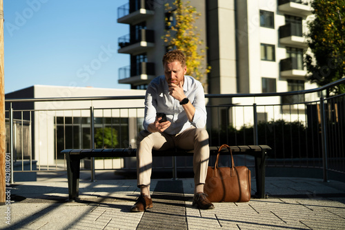 Young stylish businessman sitting on the beanch outdoors. Portrait of handsome man using the phone