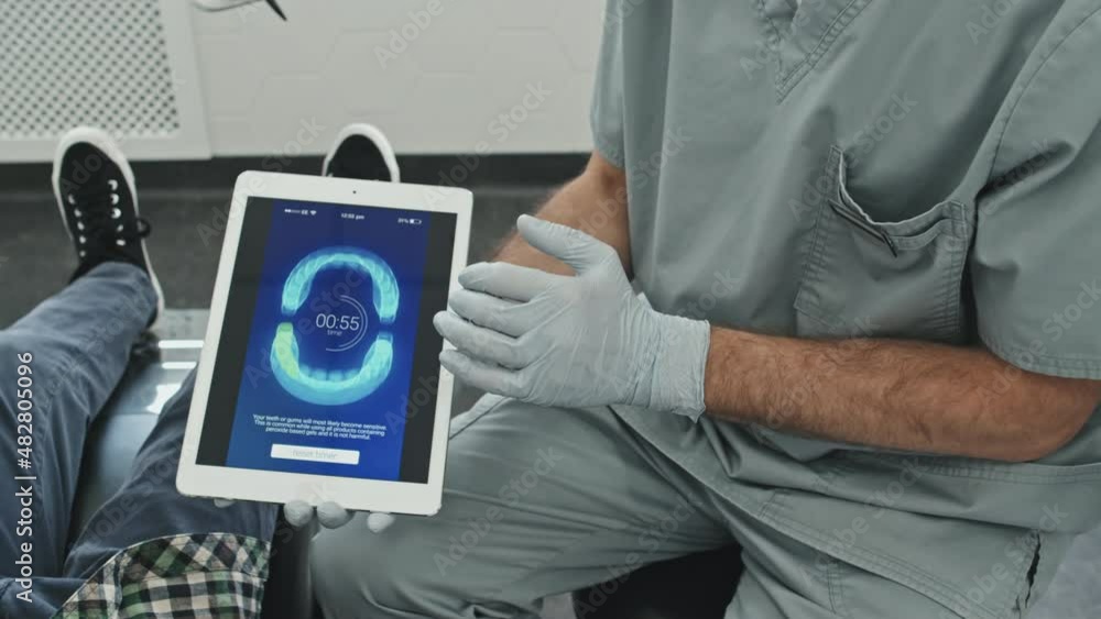 High-angle of cropped male doctor wearing scrubs and gloves, showing image of perfect bite on screen of tablet computer to curly-haired little patient sitting in dentist chair at daytime High-angle of cropped male doctor wearing scrubs and gloves, showing image of perfect bite on screen of tablet computer to curly-haired little patient sitting in dentist chair at daytime
