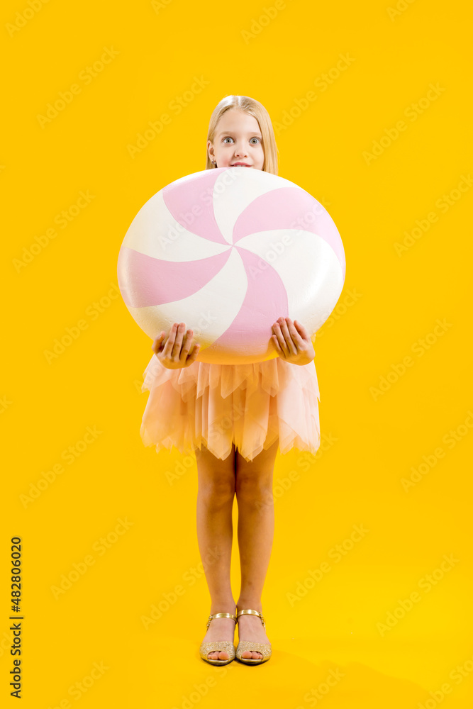 Christmas sweetness. A cute girl is holding a huge pink and white candy on a yellow background. Sweet gift. Large lollipop Christmas sweetness. A cute girl is holding a huge pink and white candy on a yellow background. Sweet gift. Large lollipop