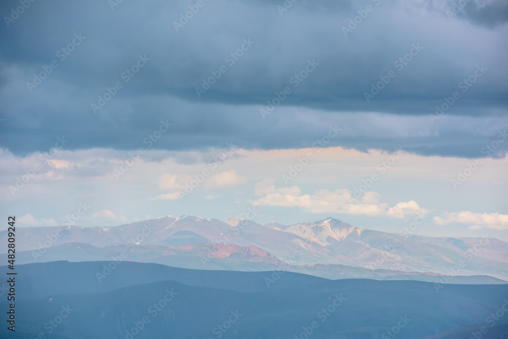 Dramatic aerial view to large multicolor mountains in haze under rain clouds at changeable weather. Scenic mountain landscape with bright sun and rainy cloudiness at same time. Motley mountain scenery