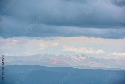 Dramatic aerial view to large multicolor mountains in haze under rain clouds at changeable weather. Scenic mountain landscape with bright sun and rainy cloudiness at same time. Motley mountain scenery