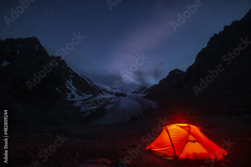 Fototapeta Naklejka Na Ścianę i Meble -  Awesome mountain landscape with vivid orange tent near large glacier tongue under clouds in night starry sky. Tent glow by orange light with view to glacier and mountains silhouettes in starry night.