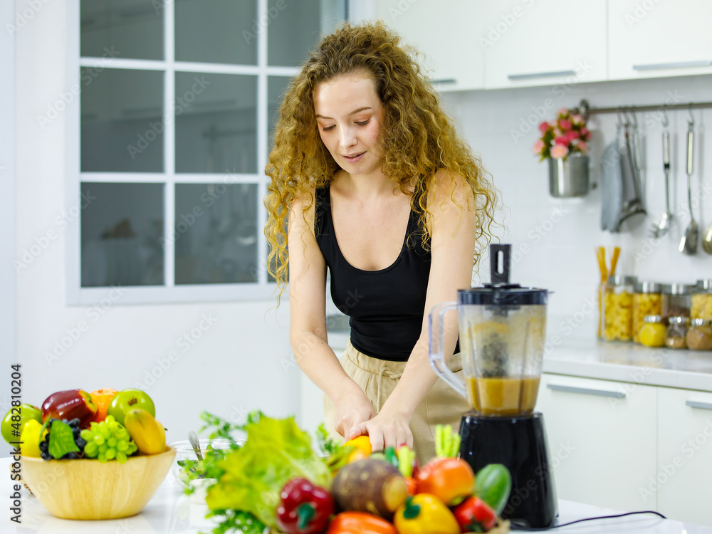Happy healthy curly hairstyle young sexy Caucasian pregnancy mother model in casual outfit holding showing sliced orange dancing around kitchen using blender mixing blended fruit and veggie juice