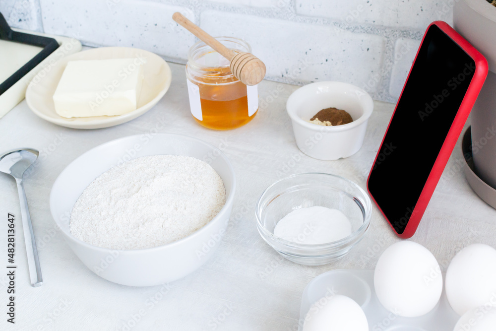 The concept of online lessons in the preparation of cookies. A smartphone in a red case and ingredients are on the table. Everything is ready for mixing future baking. Horizontal Orientation The concept of online lessons in the preparation of cookies. A smartphone in a red case and ingredients are on the table. Everything is ready for mixing future baking. Horizontal Orientation