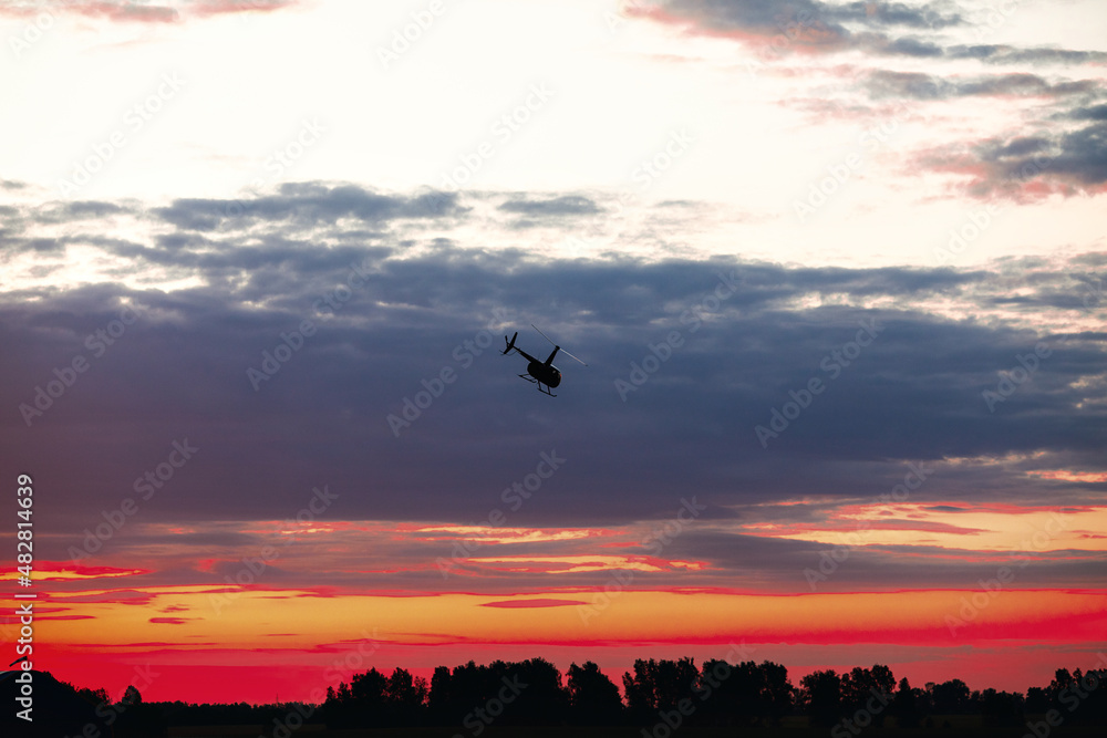 Silhouette of a chopper high in the evening sky at sundown seen from below, the line of the forest in contre-jour.