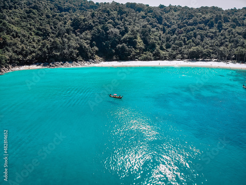 Drone view of the lagoon: narrow strip of the white sand, clear blue water and a small boat, green hills covered with tropical forest; paradise concept.