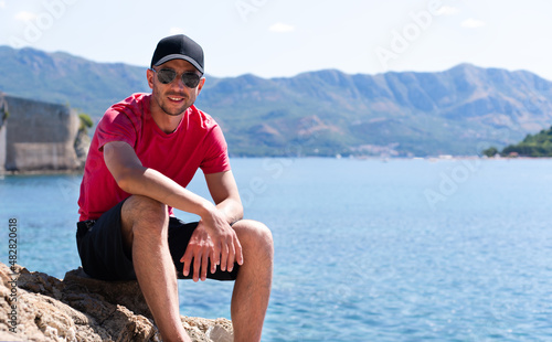 Smiling man tourist in a cap rests on the rocks on the seashore. Boka Kotorska bay in Montenegro. Copy space.