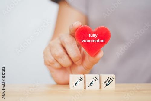 senior's hand holding red heart shape with fully vaccinated word over three syringe icon on wood cubes due to spread of corona virus, population, social or herd immunity concept