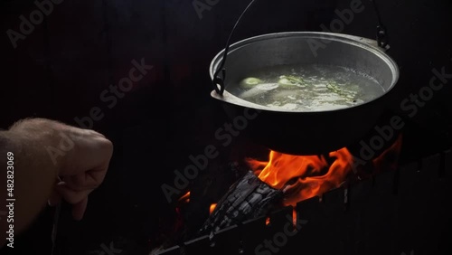 Man's hand turns burning wood in the grill. Pot of soup hangs over the fire.