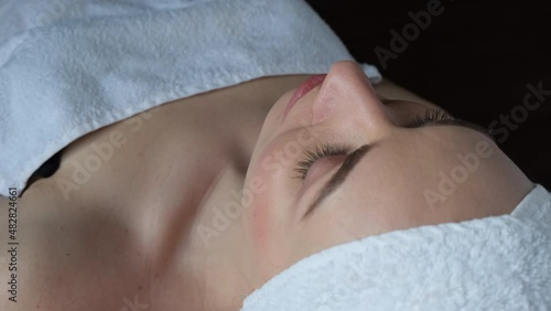 A young woman in a spa lies on a cosmetology table with a white towel on her head and perfect even soft smooth skin	  