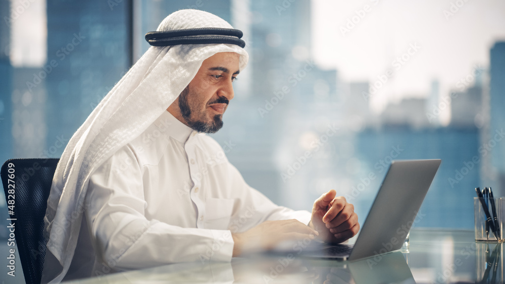 Arab Businessman in White Traditional Outfit Sitting in Office and Working on Laptop Computer. Business Manager Make Successful Investment Deal. Saudi, Emirati, Arab Businessman Concept.