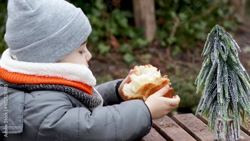 hansome 4 years old boy is eating a croisant with chochlate in puplic park, sitting at the wooden table with artificial little fir tree on it. kid in grey cap and orange furry scard. delicios street  photo