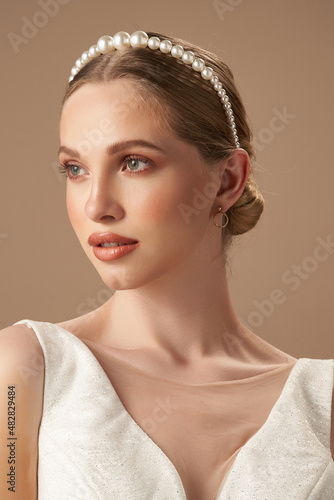 A blonde European girl in a white glitter blouse and golden earrings with charms is posing on the beige background. The girl s hair is fixed with a thin headband adorned with shiny decorative pearls.