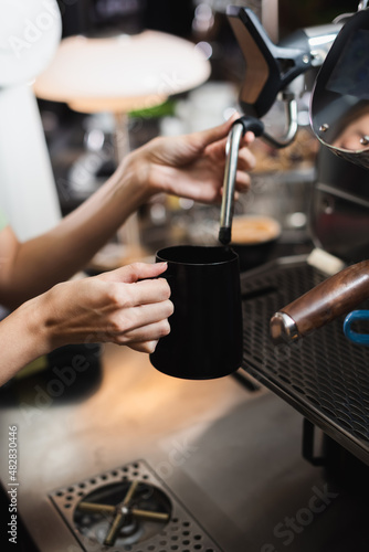 Cropped view of barista holding milk jug near steam wand of coffee machine in cafe.