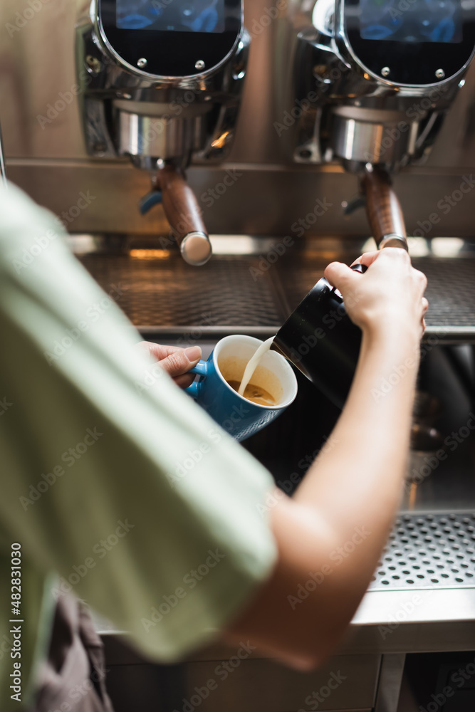 Cropped view of barista pouring milk in cup with coffee near coffee machine in cafe.