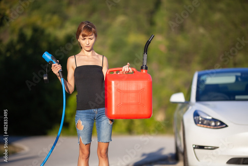 Confused girl standing next to an electric car. Holding charging cable and gassoline canister